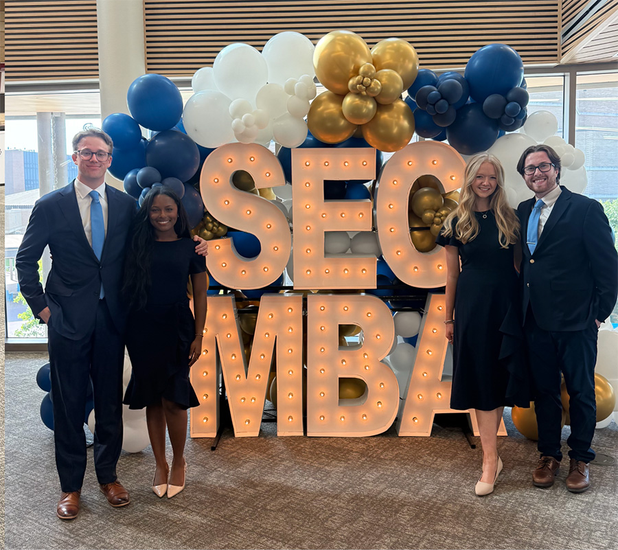 A group of four student smiling with light up SEC MBA behind them.
